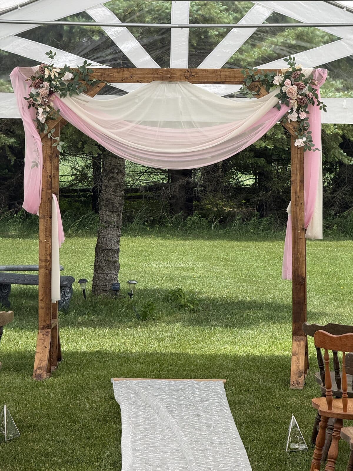 Wooden ceremony arch draped in rose fabric with florals at the end of a lace aisle runner