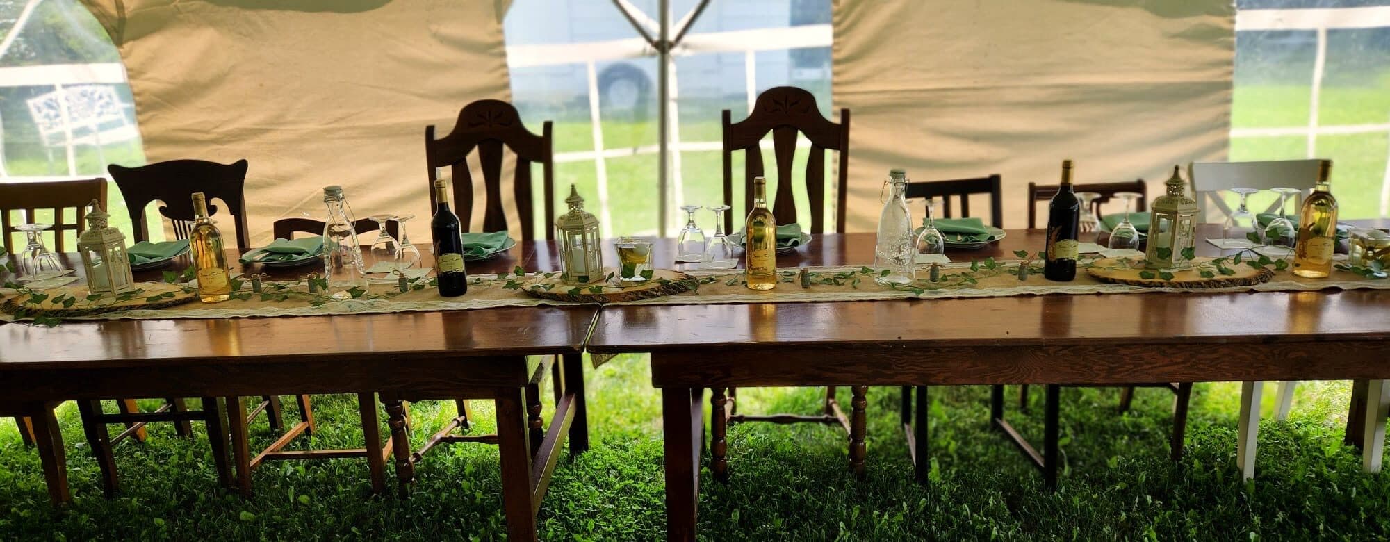Head table dressed with ivy runner, lanterns, wine bottles, and mismatched wooden chairs inside the tent